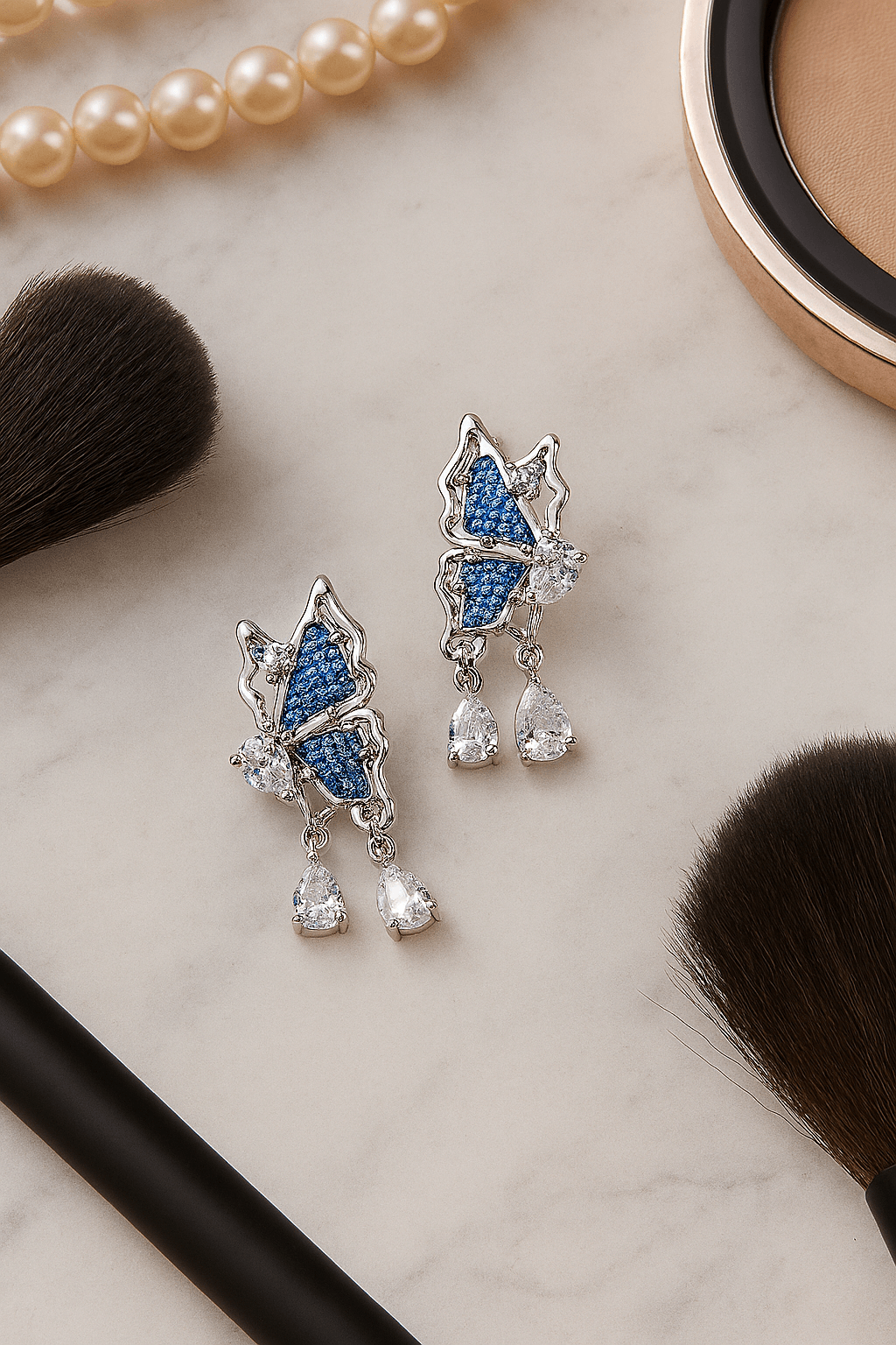 Blue Denim Butterfly Drop Earrings with Crystal Teardrop Sparkle on a marble surface with makeup tools.
