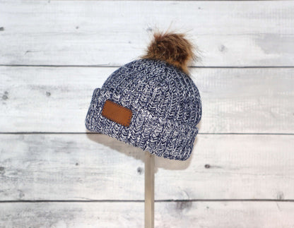 Infant Acrylic Ribbed Cuff with Pom Hat – Cozy & Purposeful displayed on a stand against a rustic wooden background.