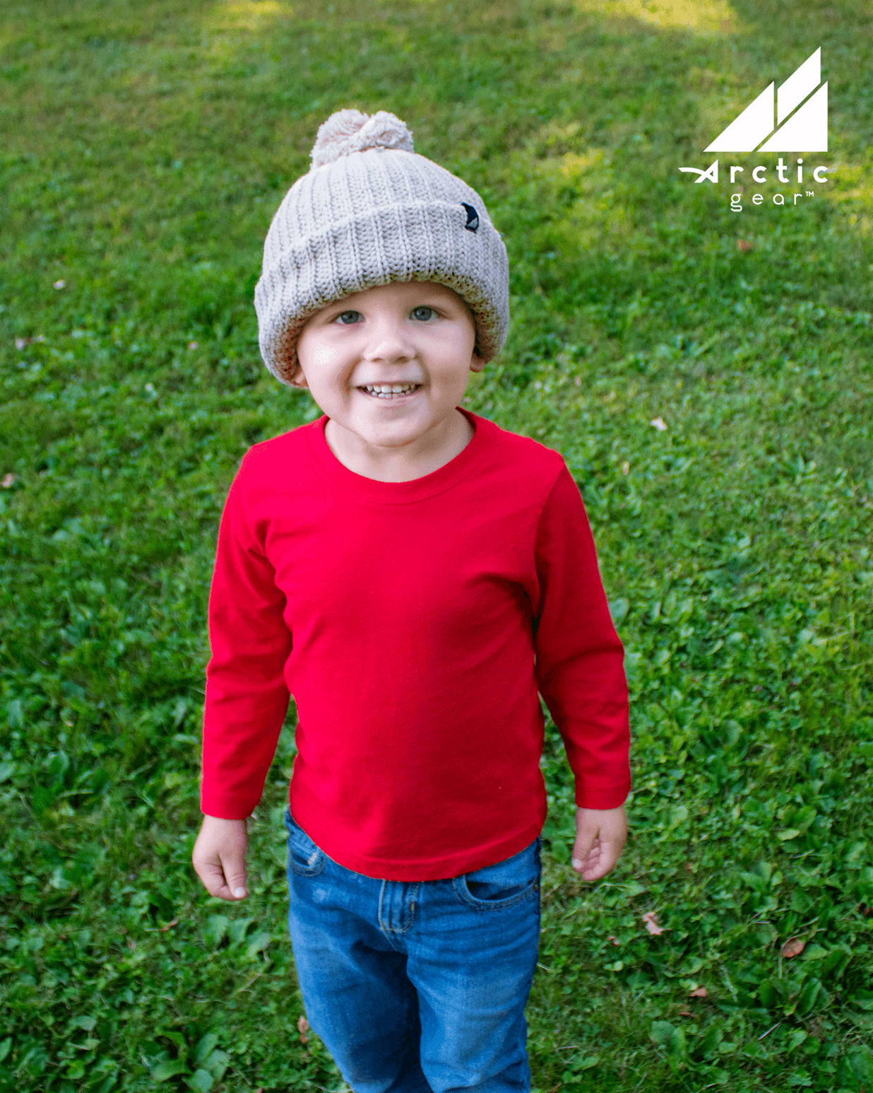 Child Acrylic Ribbed Cuff Hat with Pom – Cozy, Playful, Purposeful worn by a smiling child in a red shirt.