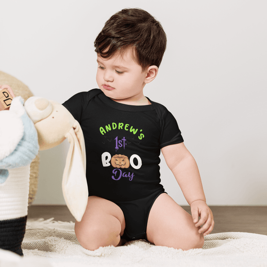 Adorable baby in black bodysuit with 'Andrew's 1st BOO Day' design and pumpkin graphic, sitting with stuffed bunny.