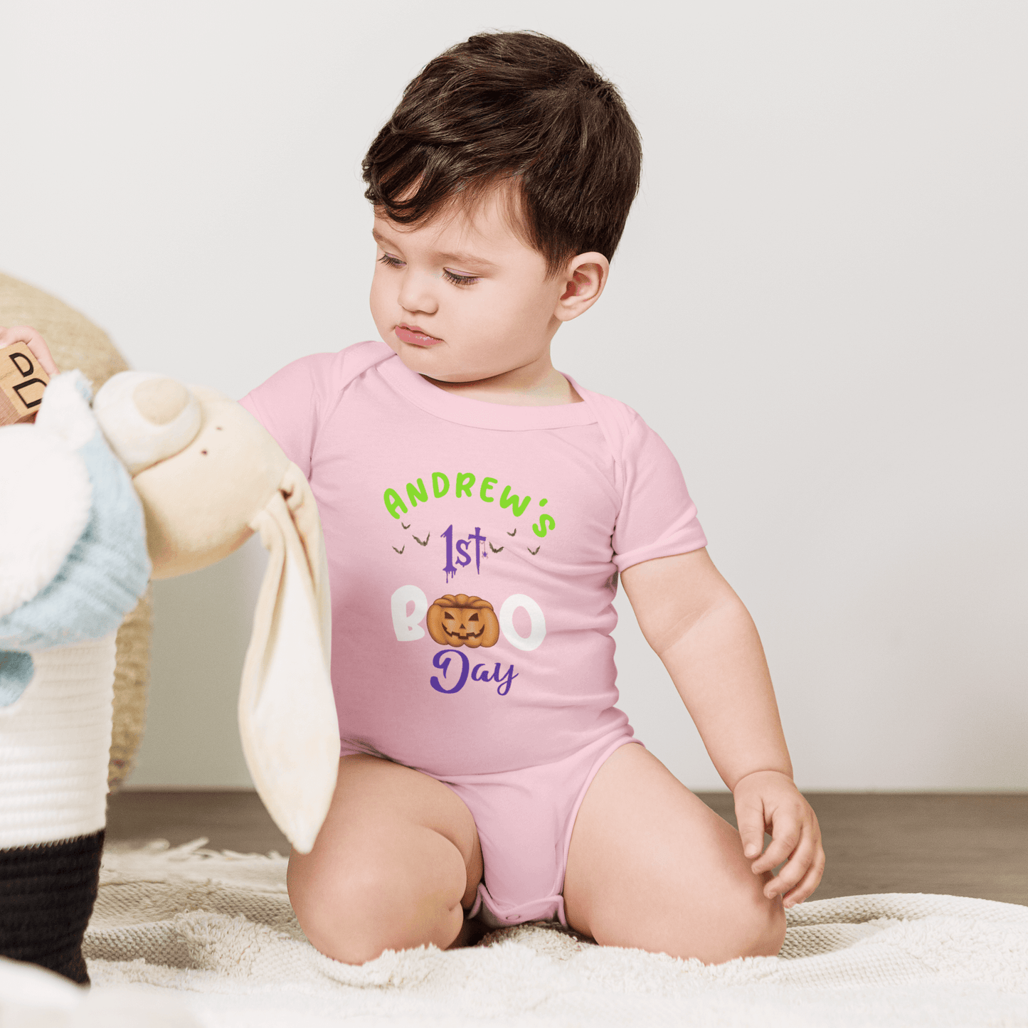 Baby in personalized pink Halloween bodysuit with '1st BOO Day' text, playing with stuffed bunny.