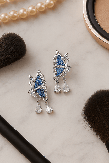 Blue Denim Butterfly Drop Earrings with Crystal Teardrop Sparkle on a marble surface with makeup tools.