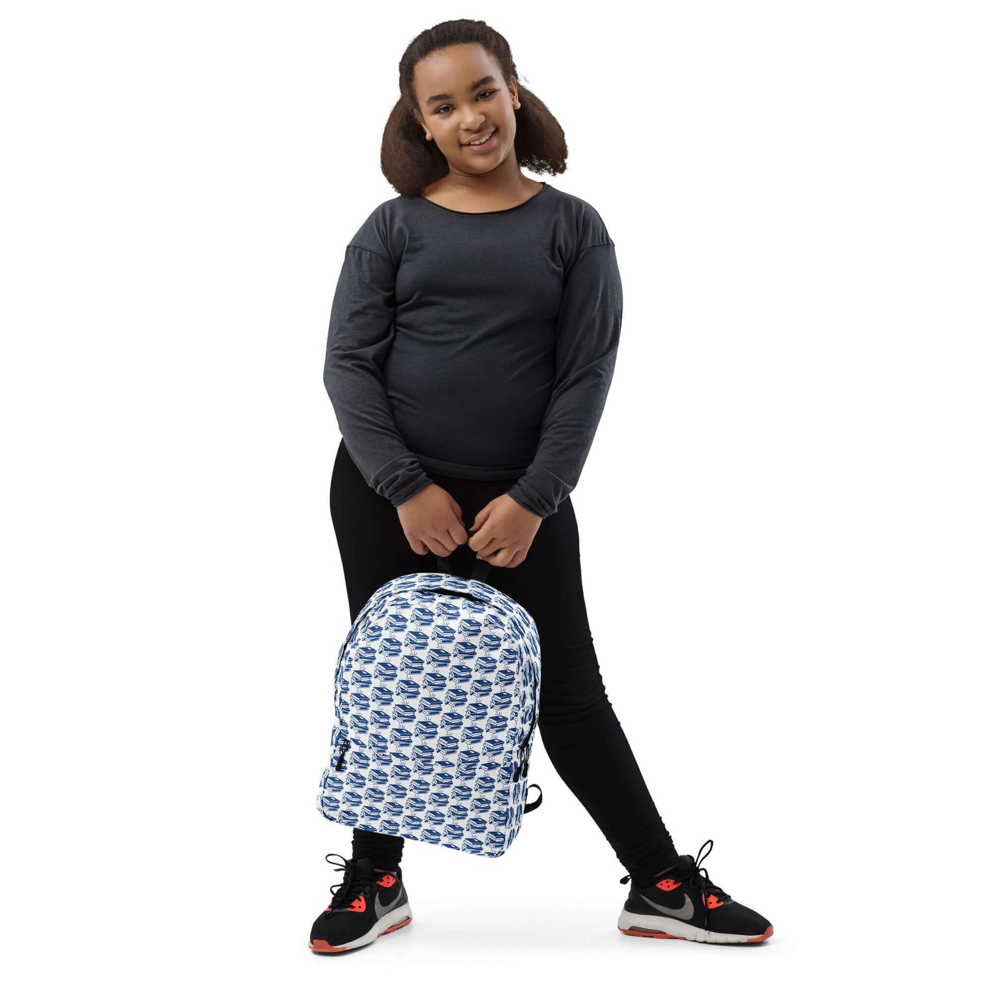 Young girl wearing a black top with a geometric book-pattern backpack, smiling happily.