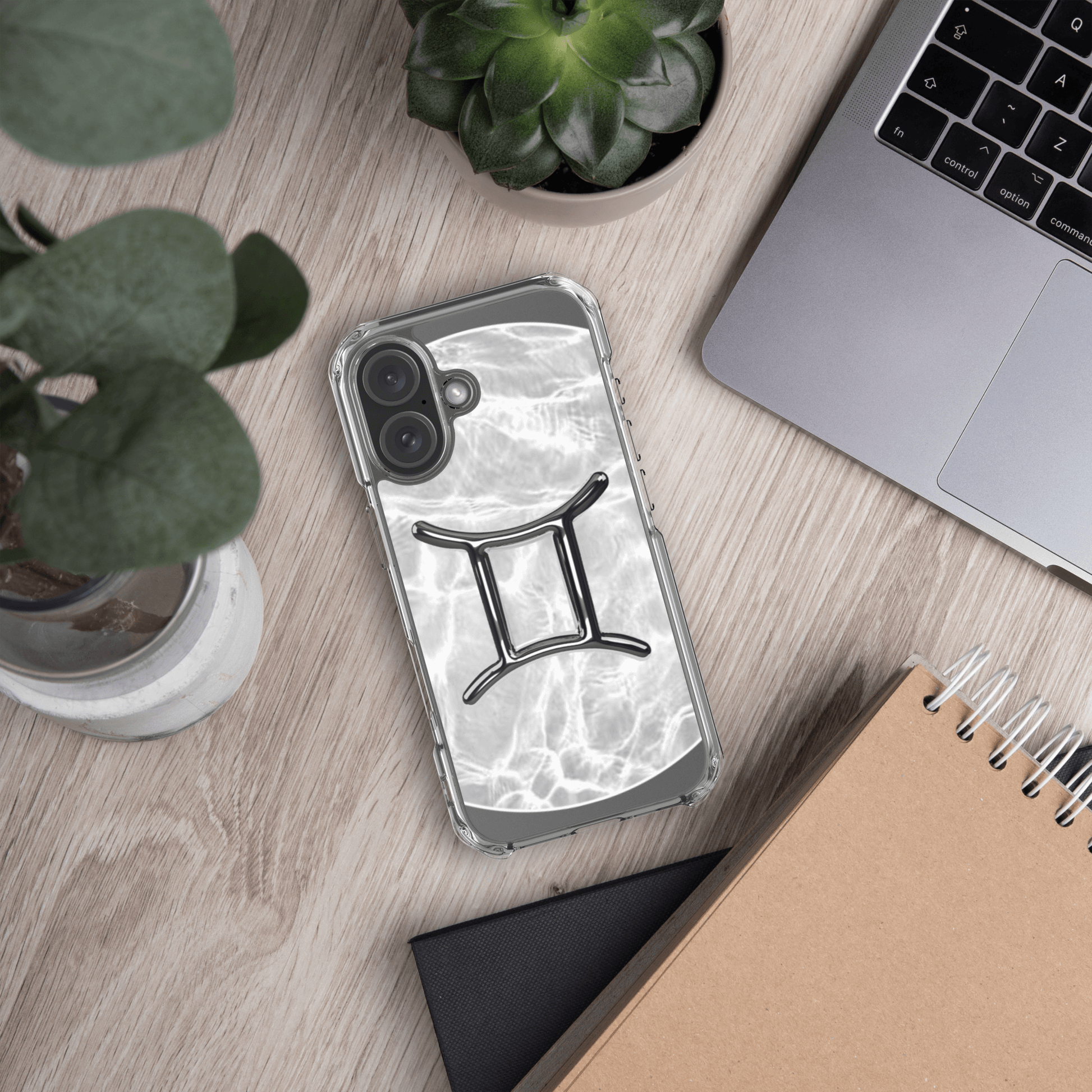Zodiac Gemini Magsafe clear iPhone case with metallic design on a wooden desk with plants and a laptop.