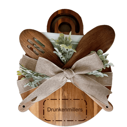 Personalized wooden cheese board with kitchen utensils and greenery, engraved with 'Drunkenmillers'.