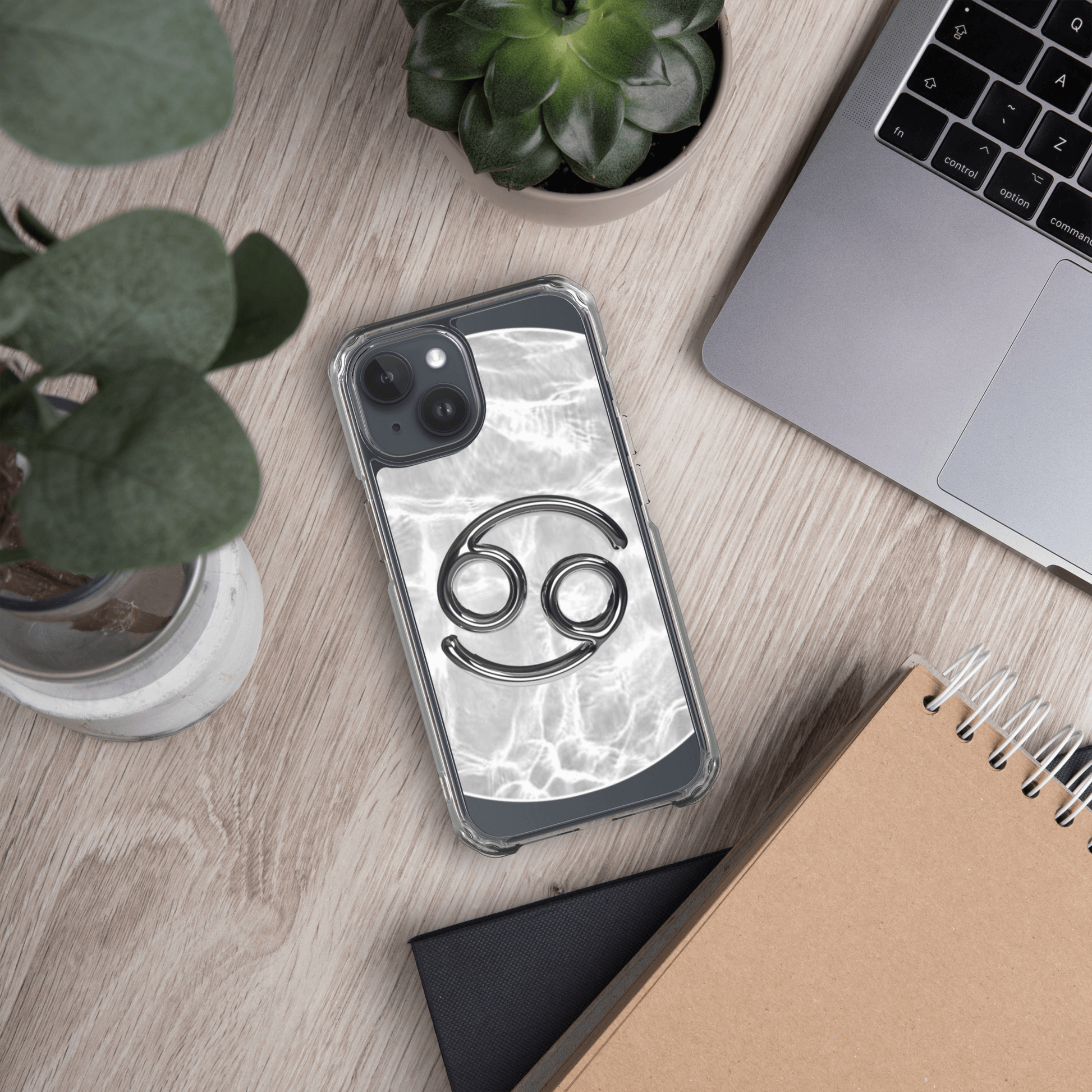 Clear iPhone case with Zodiac Cancer design on a marble surface, surrounded by plants and a laptop.