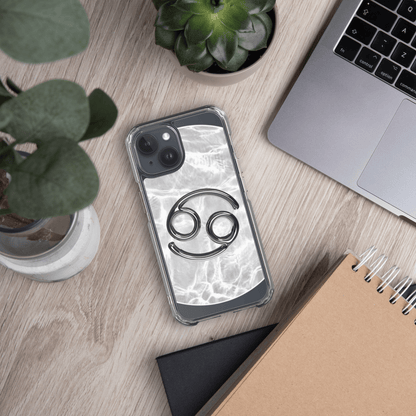 Clear iPhone case with Zodiac Cancer design on a marble surface, surrounded by plants and a laptop.